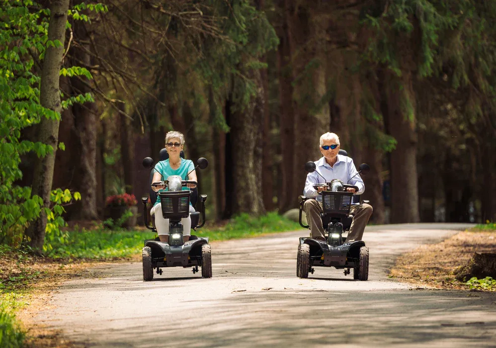 Two people riding mobility scooters along a tree-lined road